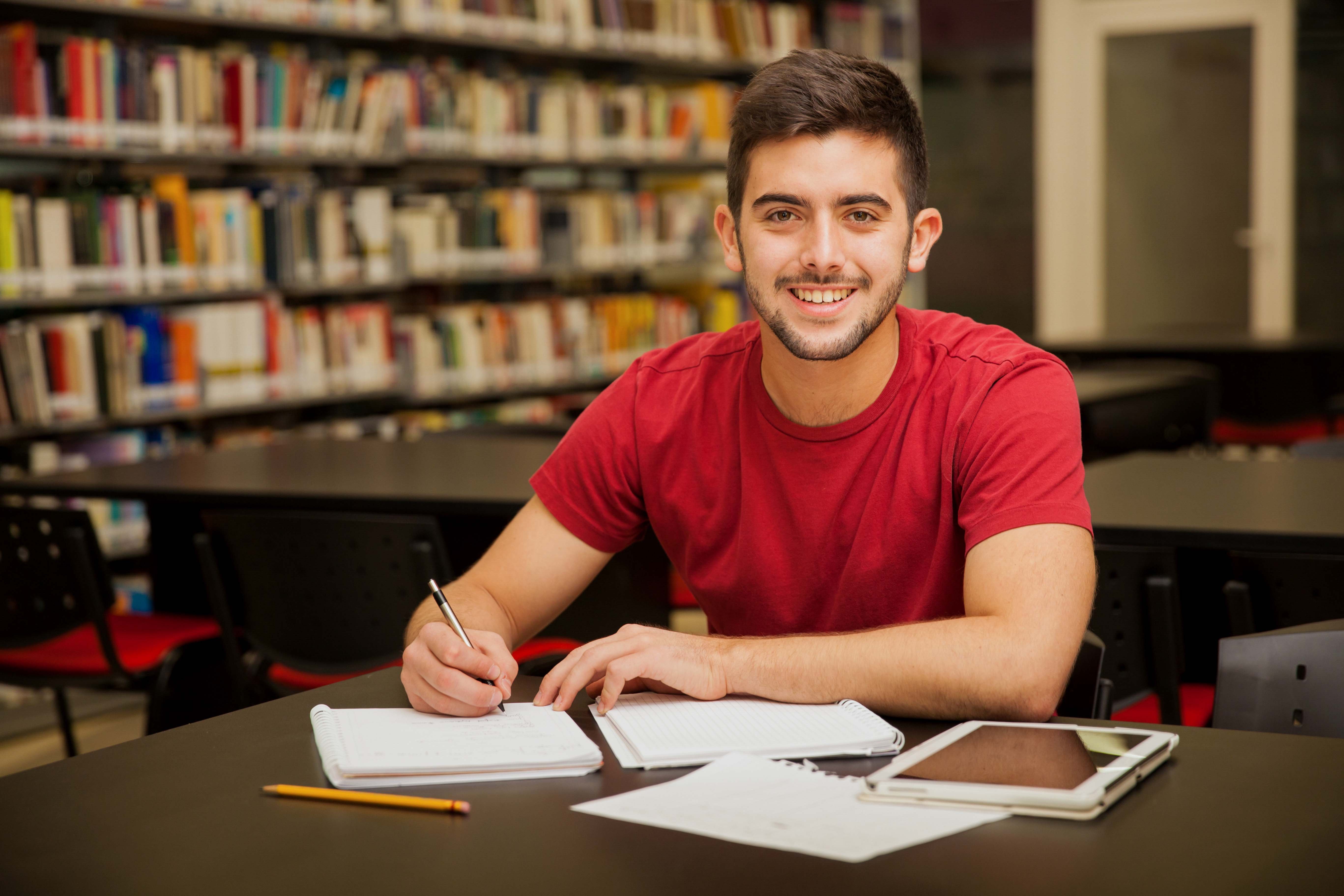 attractive-male-university-student-doing-some-homework-school-library-smiling
